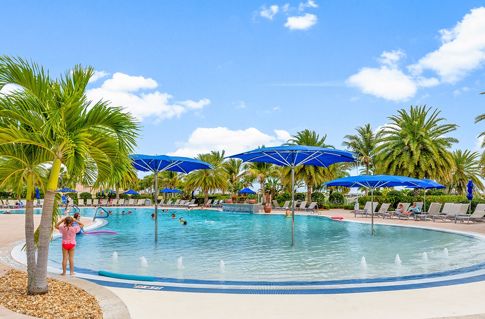 Resort-style pool with fountains, in-water loungers, and blue umbrellas surrounded by palm trees under a sunny sky.