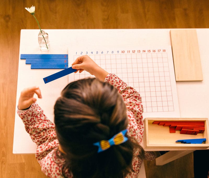 A young girl doing school work using blue measuring tools over a large paper at the Island Village Montessori School