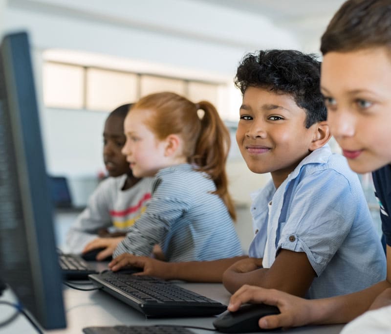 A line of kids sitting in front of computers, with one young boy looking at the camera smiling at the Sky Academy Middle School