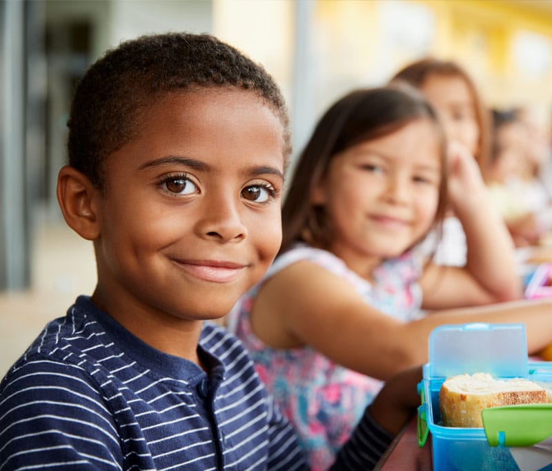 A line of Elementary school kids sitting at a table for their lunch and looking at the camera while smiling at Taylor Ranch Elementary school