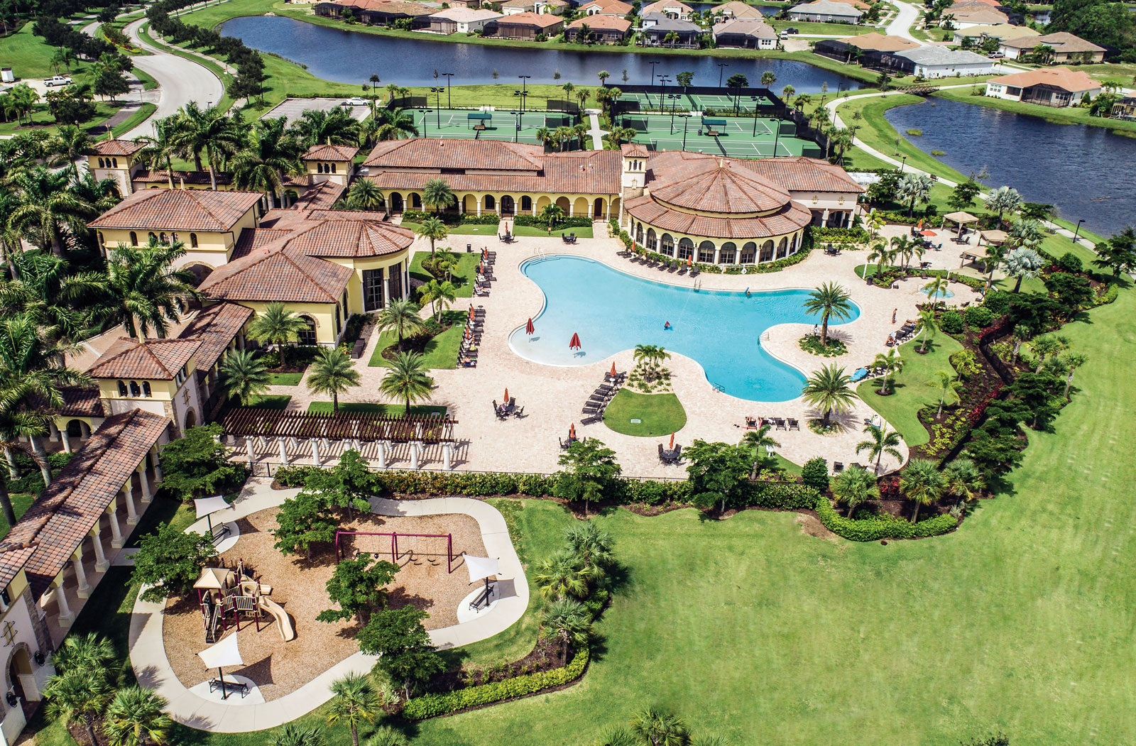 Aerial view of a luxury community with a large resort-style pool, clubhouse, tennis courts, playground, and surrounding lakes and homes.