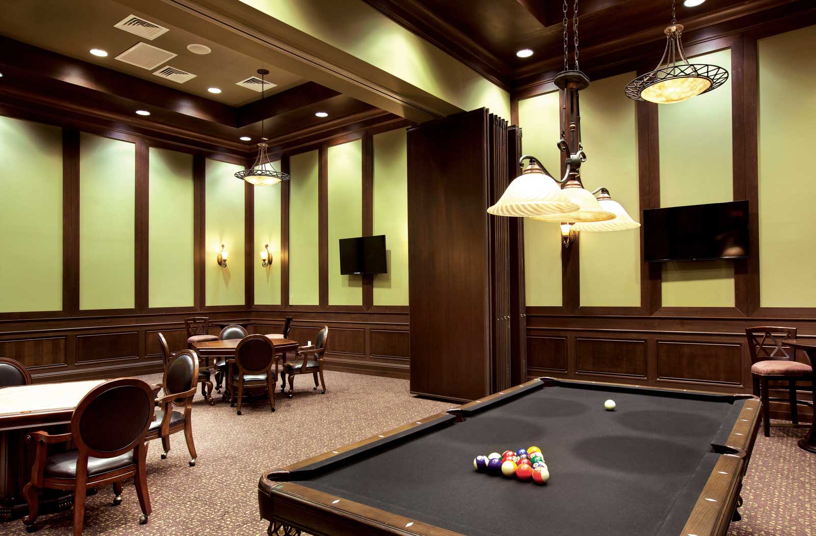 Elegant game room with a black pool table, card tables, wood-paneled walls, and hanging pendant lights, creating a classic clubhouse atmosphere.