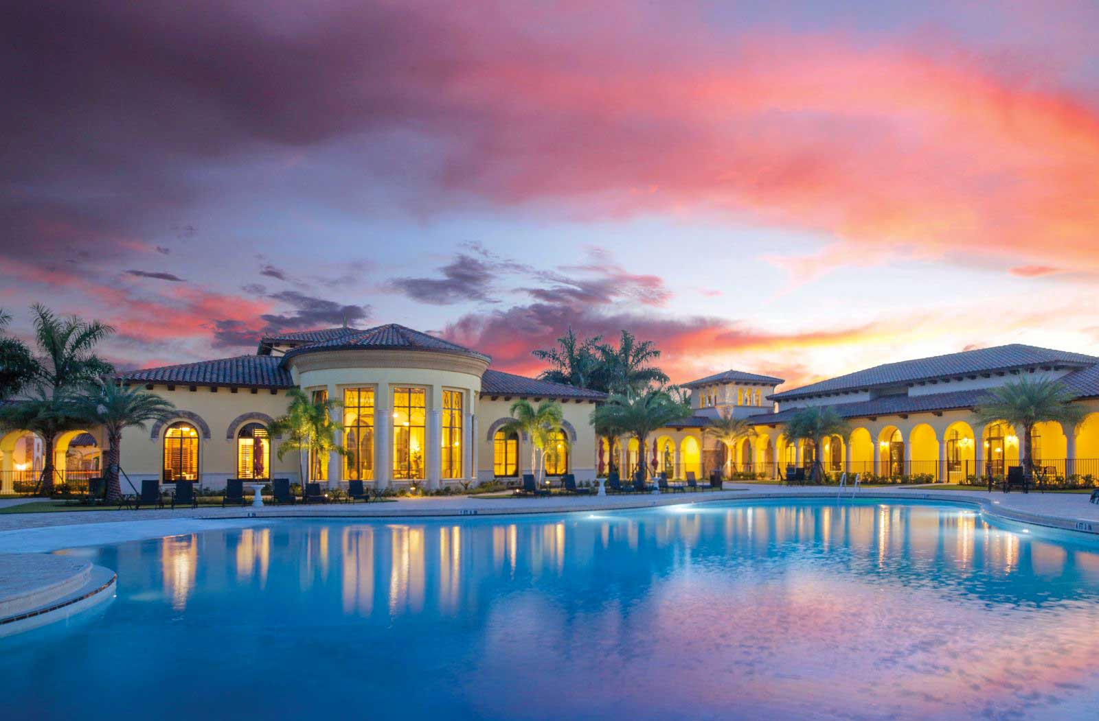 Luxurious clubhouse with arched windows and palm trees reflected in a large pool at sunset, under a vibrant sky.