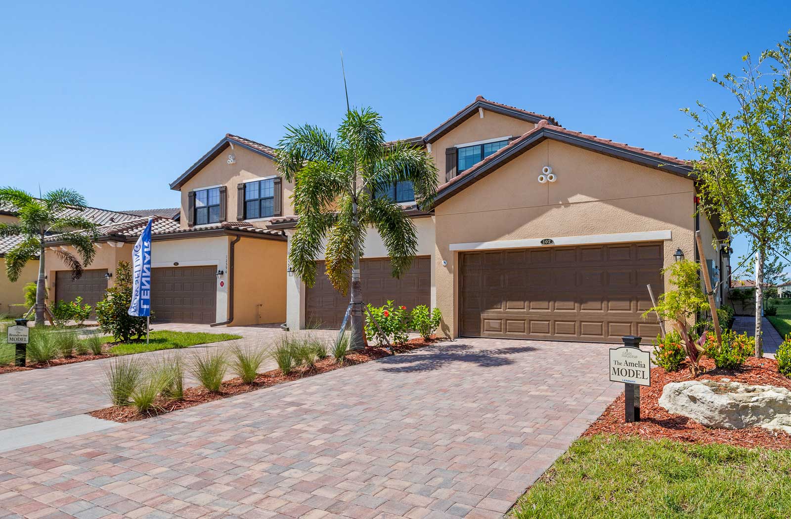 Two-story model townhome with a tile roof, brown garage doors, and tropical landscaping, featuring a sign that reads “The Amelia Model.”
