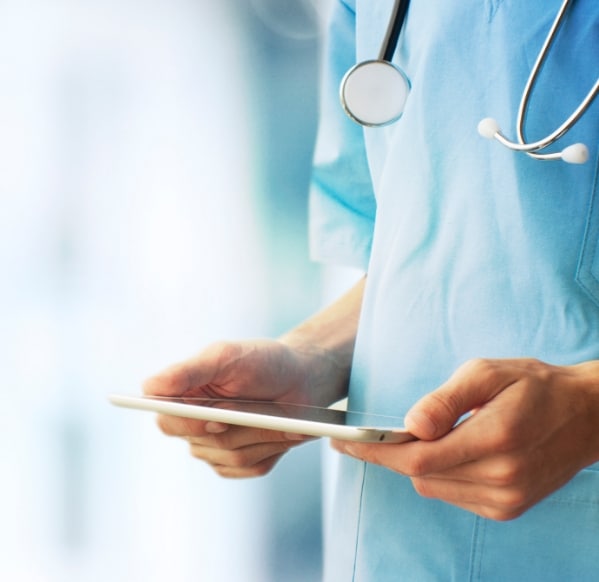 A torso shot of a health care professional in blue scrubs holding an ipad with their stethoscope hanging over their shoulders