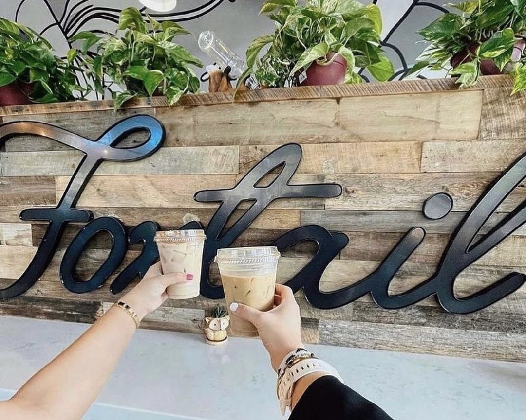 Two arms extended, cheersing their ice coffee beverages in front of the Foxtail Coffee store sign