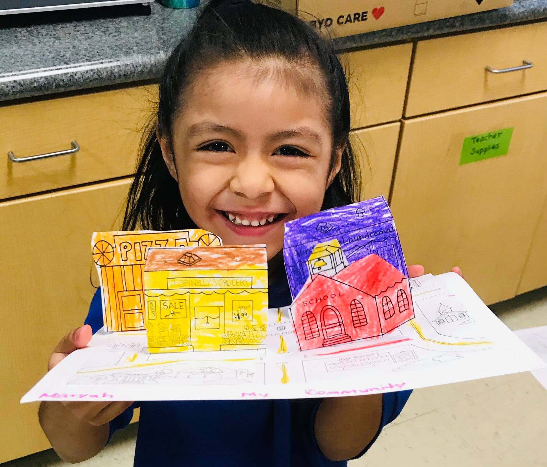 A smiling elementry school girl holding up a 3D art piece of an orange, yellow, purple and red house at the College Preparatory Academy at Wellen Park