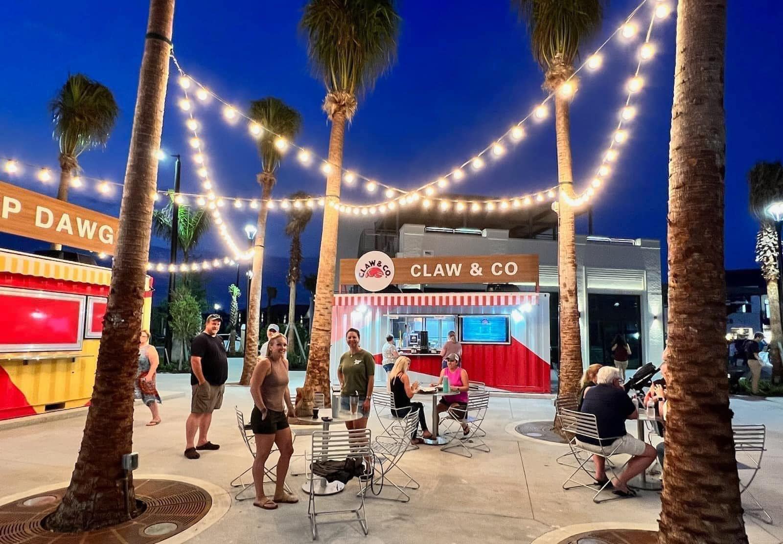 A waterfront yard with food and drink stalls, with fairy lights hung up from the surrounding palm trees