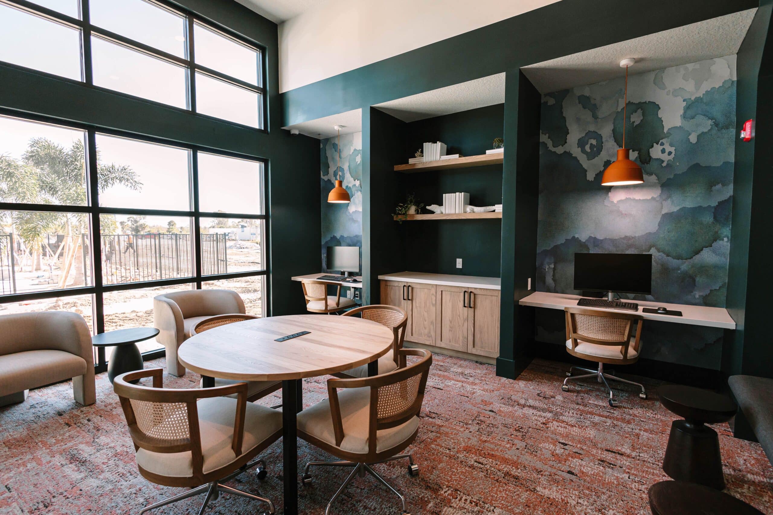 A private lounge room with a central round table for 4, a work space with a computer against the wall and floor to ceiling paneled windows with a view of the community