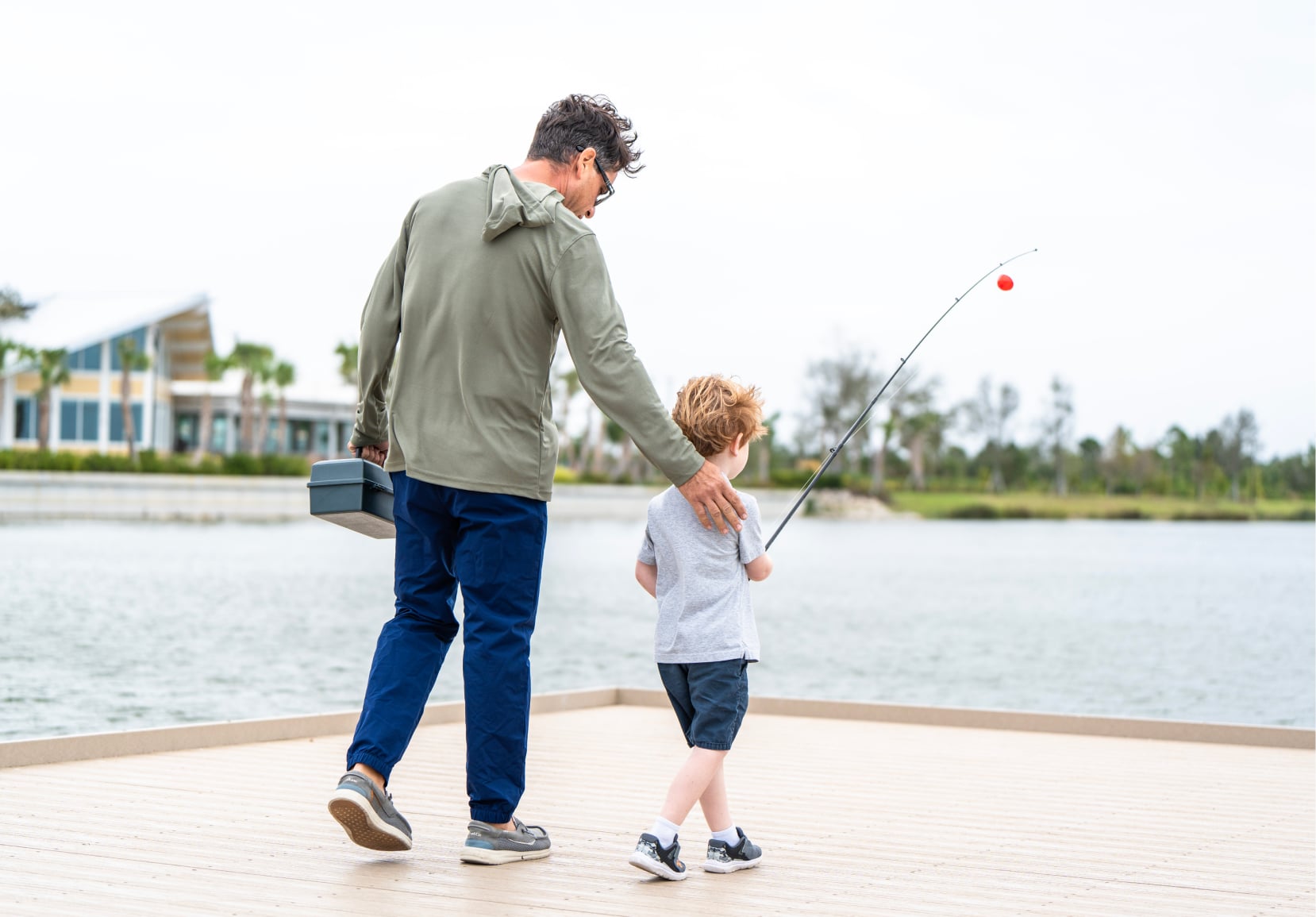 A young boy holding a fishing rod with his father approaching the edge of a dock, with houses on the waterfront in the background