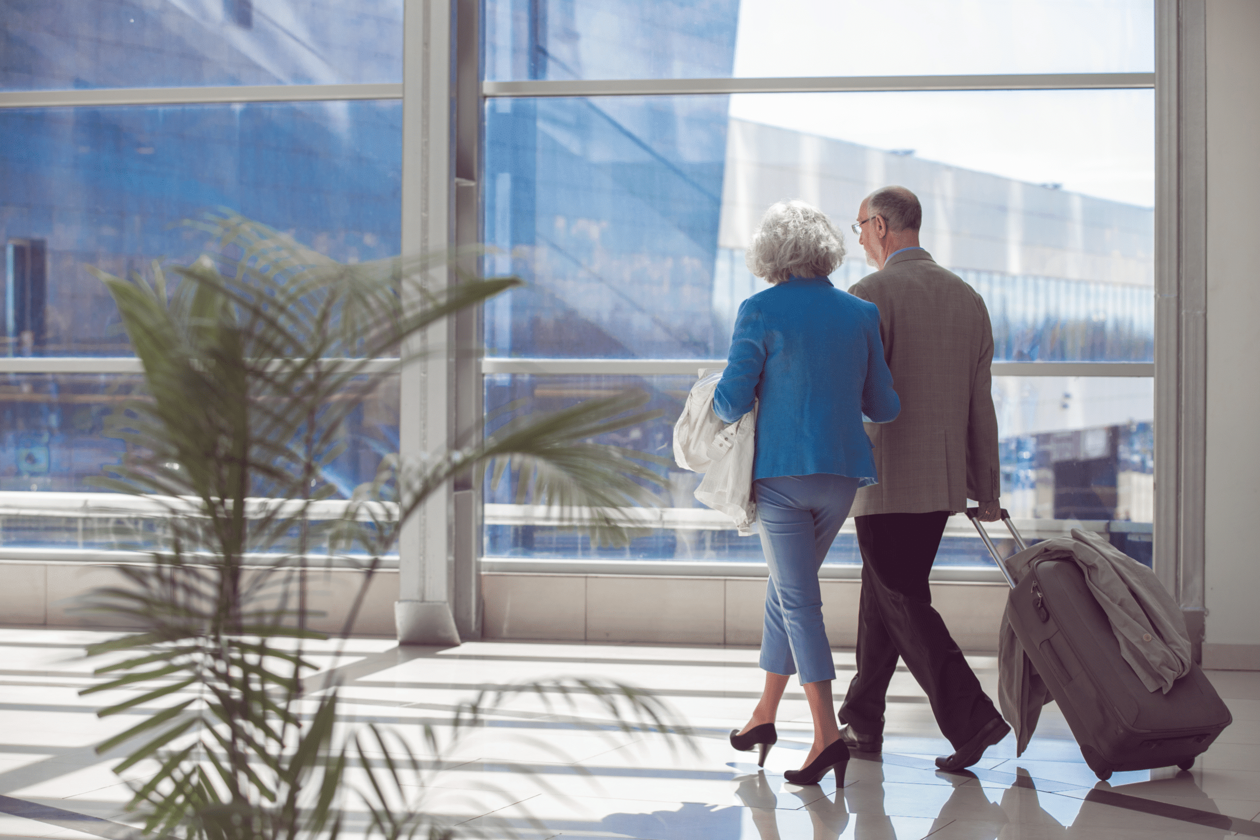 A senior couple walking down an airport hallway with the man rolling a suitcase behind him