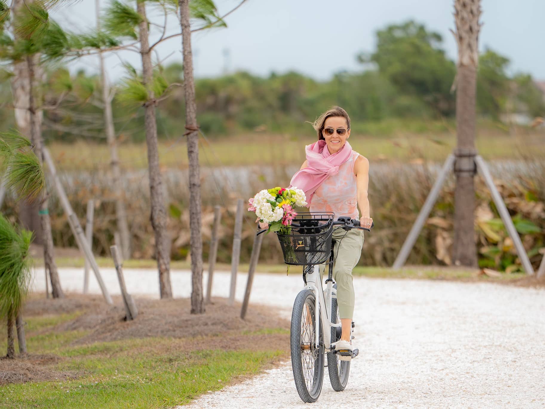 Woman riding a bike on a scenic path, smiling and wearing sunglasses and a pink scarf, with flowers in the front basket.
