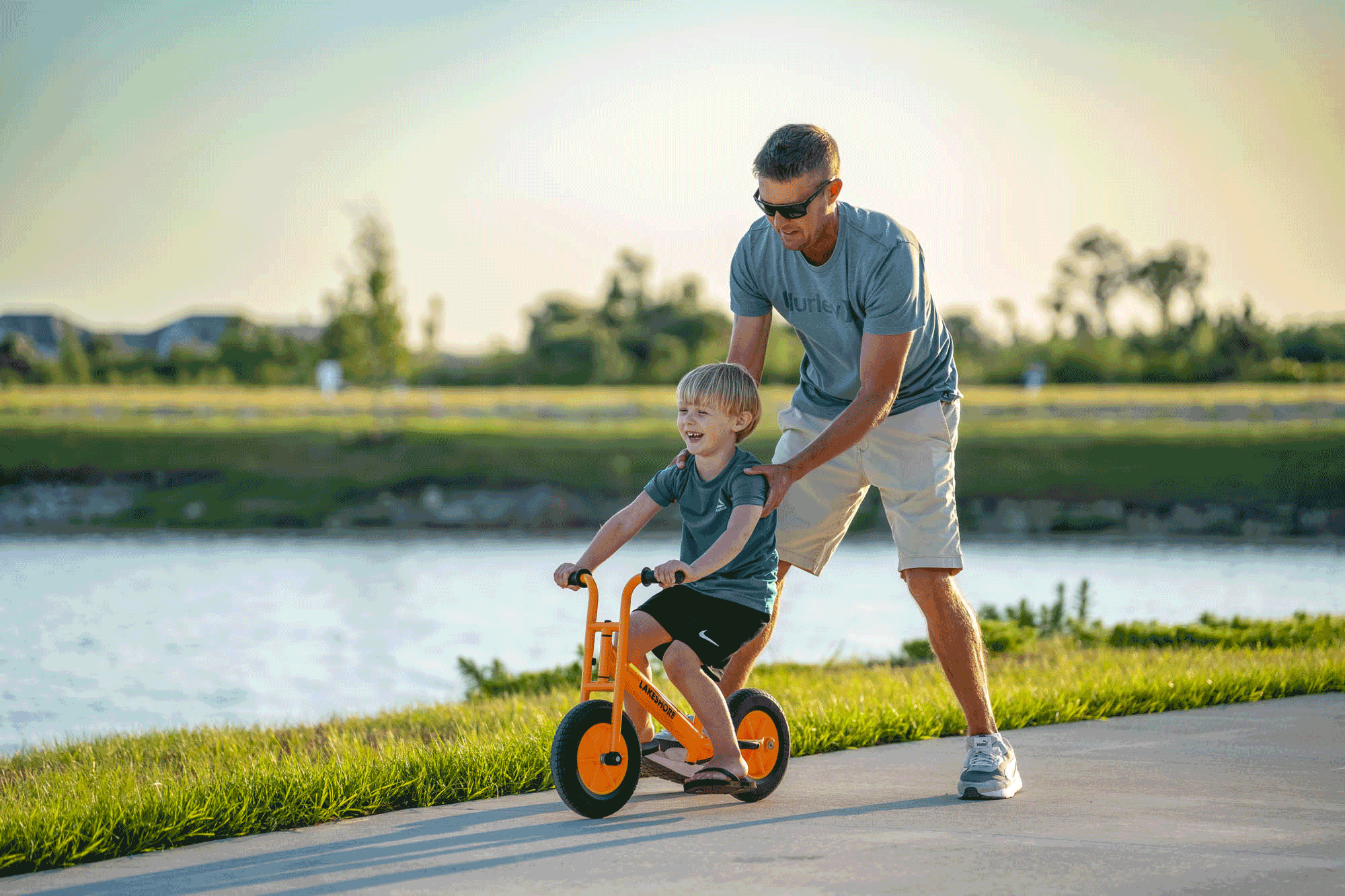 A man helping a young boy ride an orange bike on a sunny path near a pond.