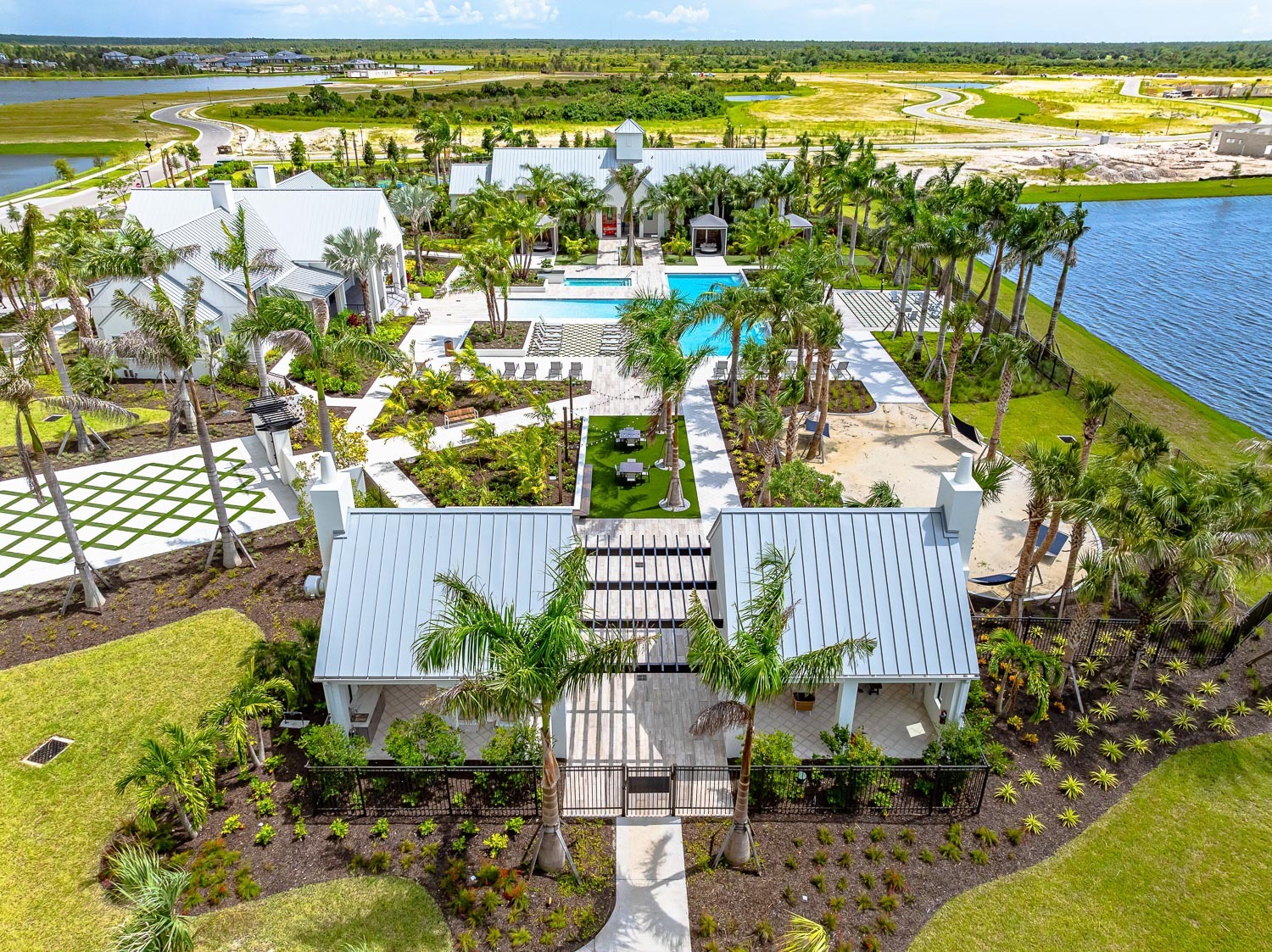 An aerial view of the Everly at Wellen pool area with a covered lounge featuring a kitchen, dining tables, a sand pit with hamocks, all leading up to a pool in the center of several lounging chairs, palm trees and other amenity buildings