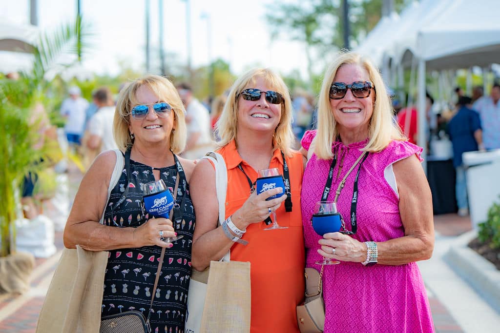 Three women smiling for a picture at an outdoor event, holding covered wine glasses