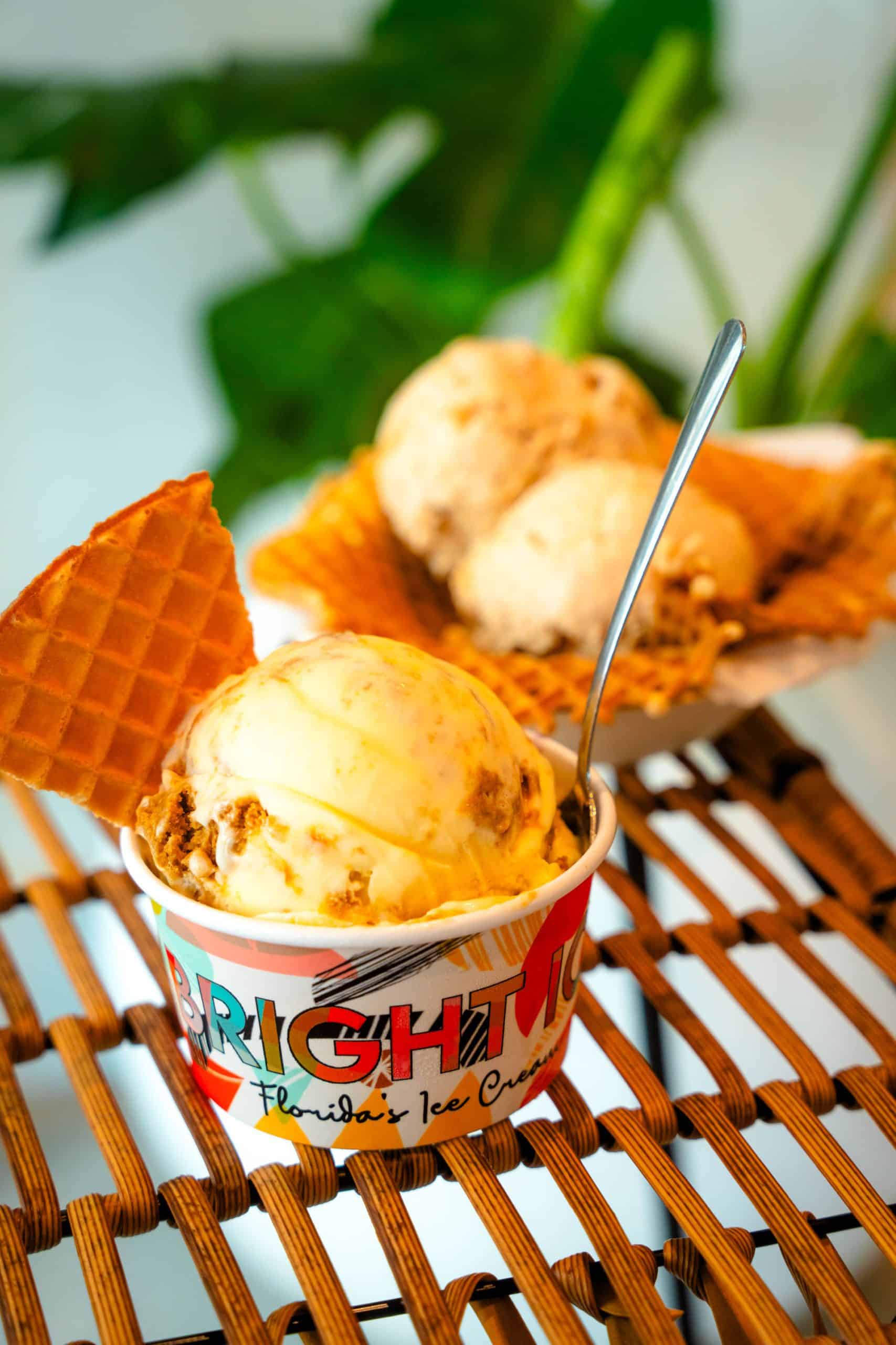 A cup of ice cream with a waffle triangle stuck in it, next to two scoops of ice cream in a waffle bowl all on a basket weave table